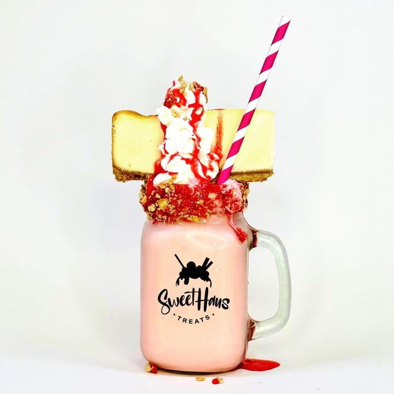 A Sweethaus Treats strawberry milkshake in a mason jar, topped with cheesecake slices, whipped cream, and strawberry drizzle.