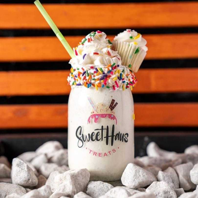 A Sweethaus Treats milkshake with whipped cream, sprinkles, a cupcake, and a green straw.